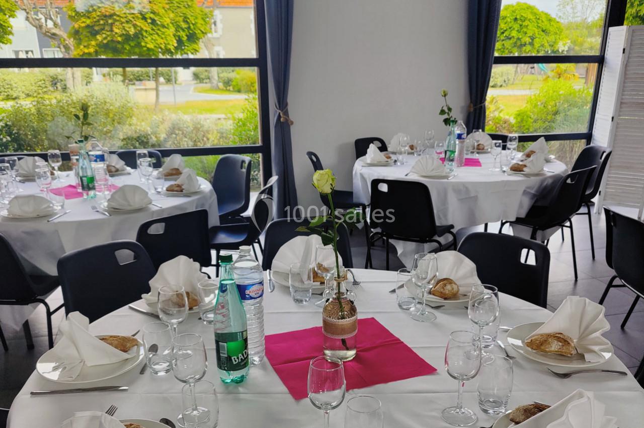 Salle de réception avec tables rondes dressées, nappes blanches, serviettes pliées, verres, et décoration florale.
