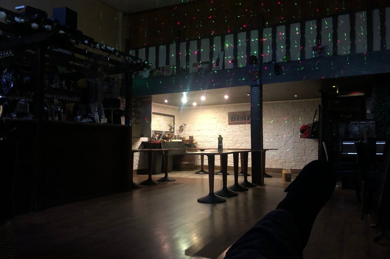 Salle sombre avec tables empilées, bar à gauche et lumières colorées projetées sur les murs et le plafond.