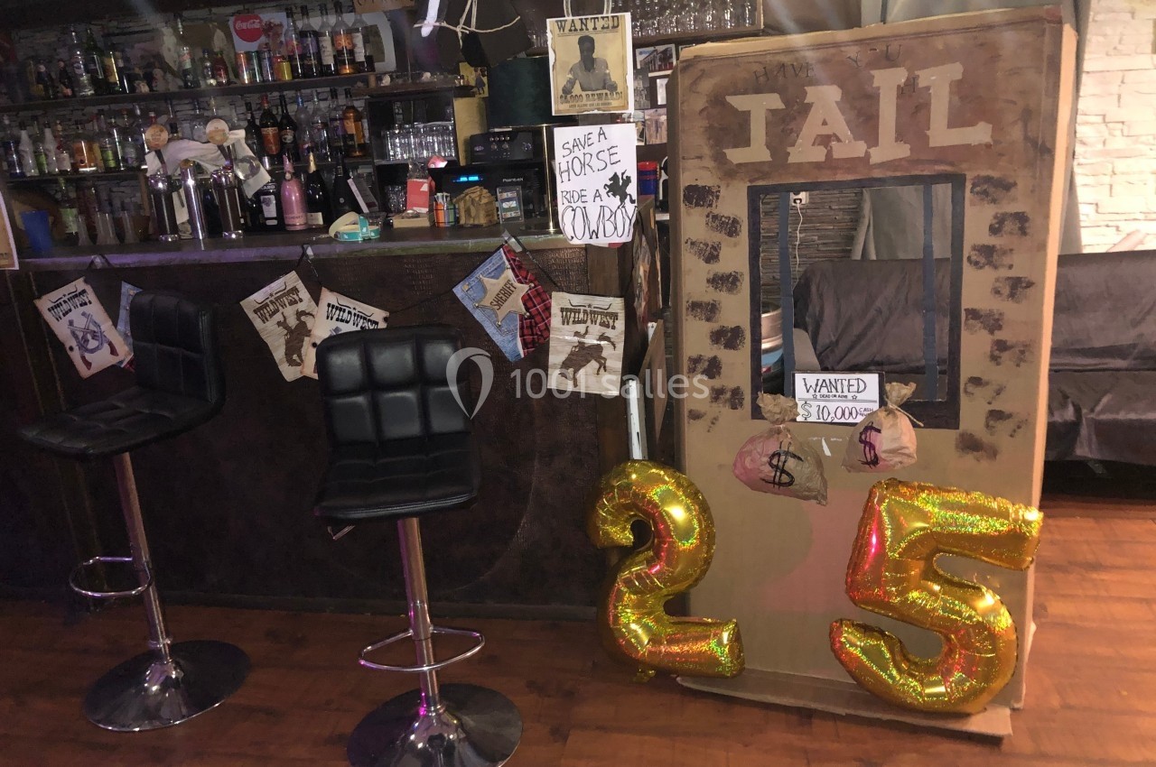 Décor de bar sur le thème du Far West avec une fausse cellule de prison, affiches ’Wanted’ et ballons dorés ’25’.