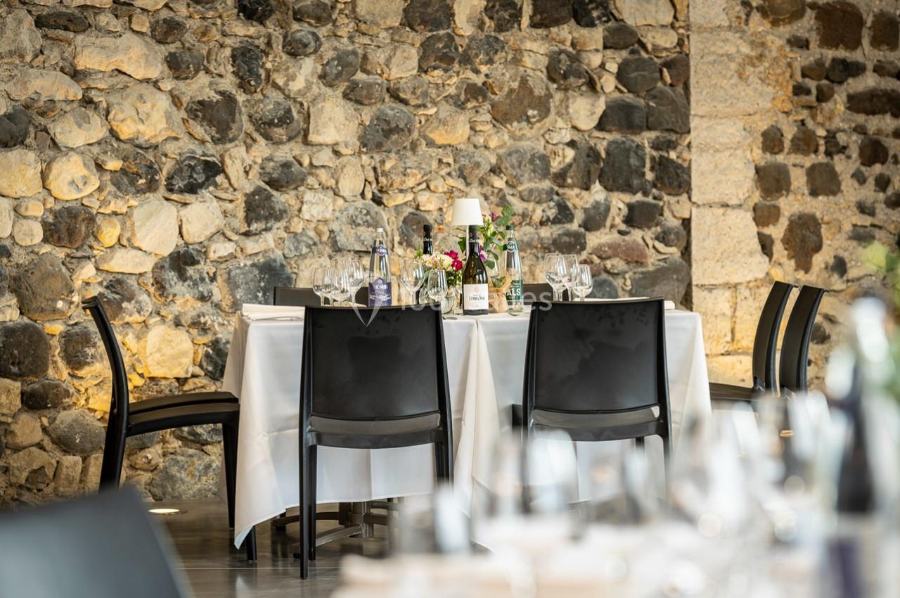 Table dressée avec nappe blanche, chaises noires, vaisselle et décoration florale, devant un mur en pierre apparente.