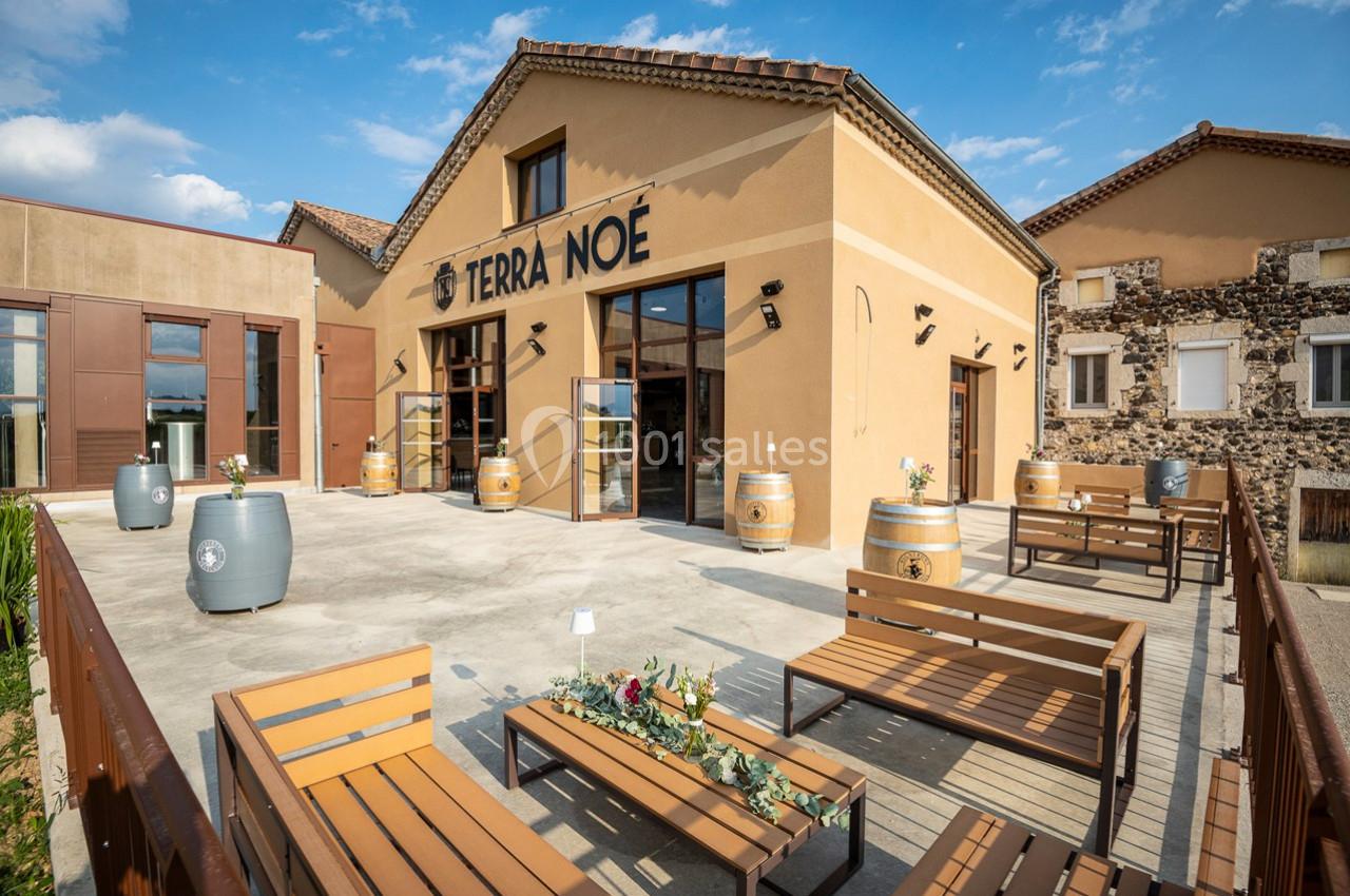 Terrasse aménagée avec bancs et tonneaux devant un bâtiment en pierre et enduit beige sous un ciel dégagé.