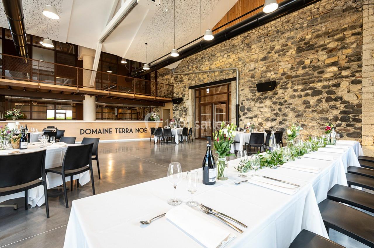 Salle de réception lumineuse avec murs en pierre, tables dressées et décorations florales.