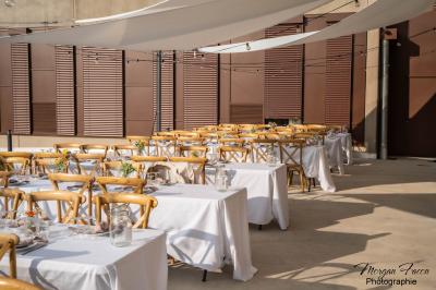 Tables dressées avec nappes blanches et chaises en bois sous des voiles d'ombrage dans une cour extérieure ensoleillée.