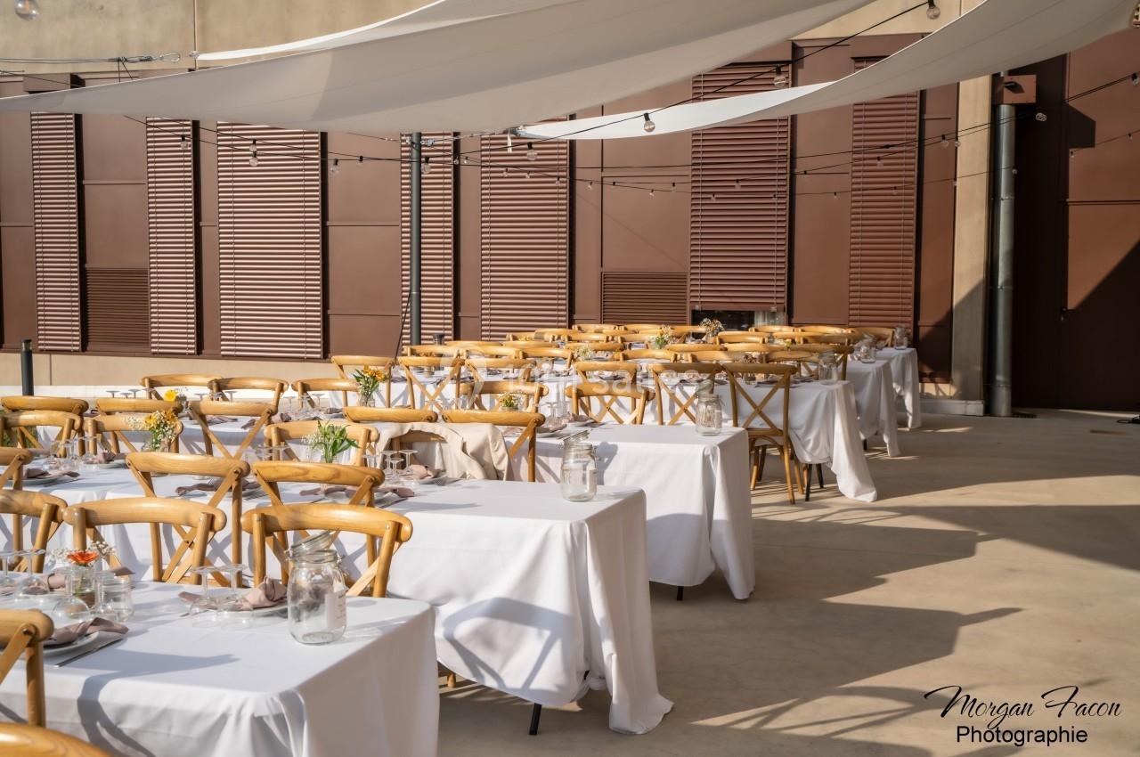 Tables dressées avec nappes blanches et chaises en bois sous des voiles d'ombrage dans une cour extérieure ensoleillée.