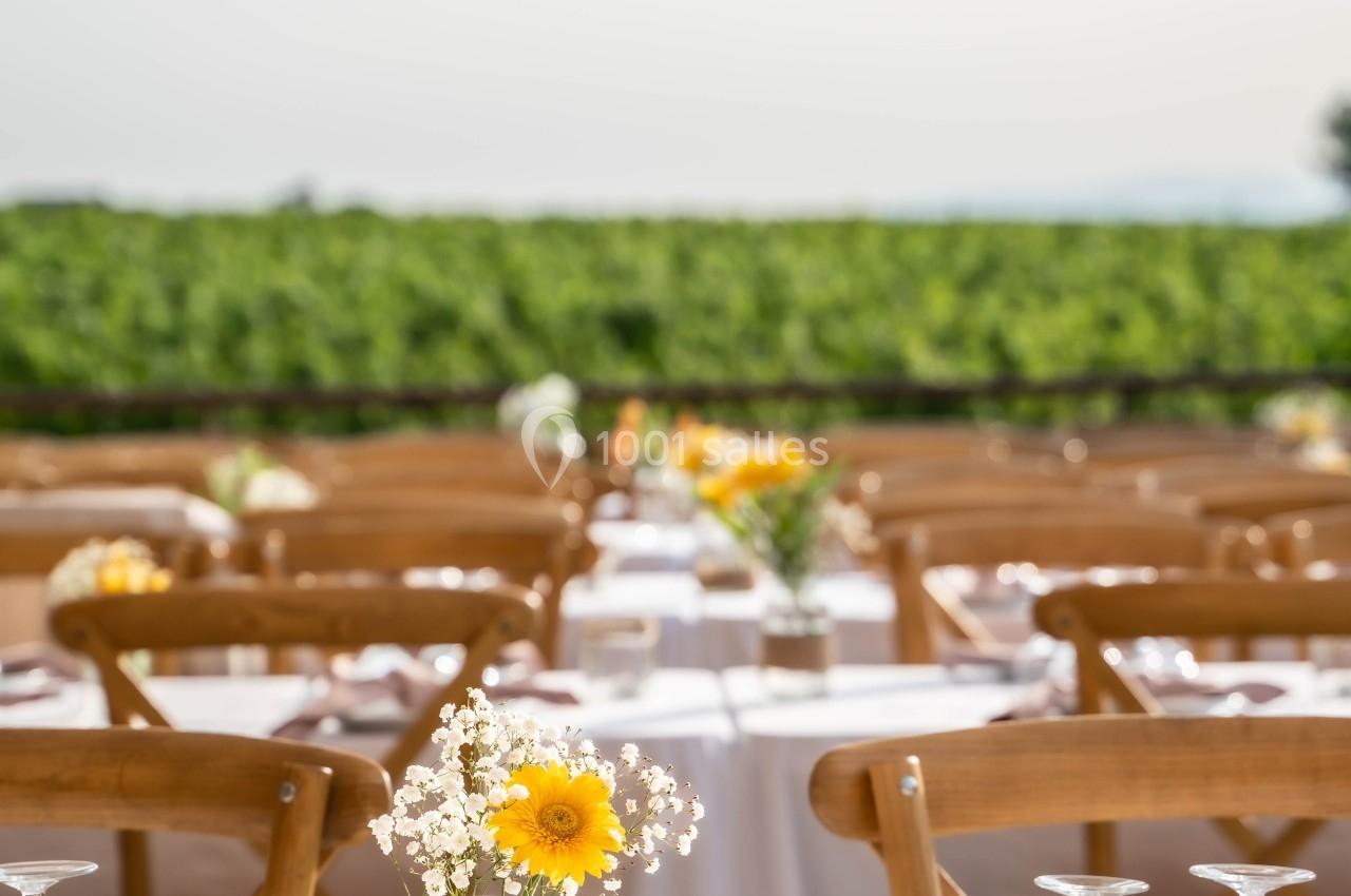Tables décorées avec des fleurs jaunes et blanches sous une tente, avec une vue sur des vignes en arrière-plan.