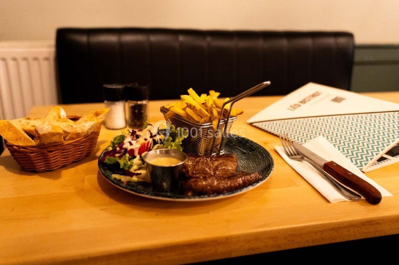 Assiette avec steak, frites, salade, sauce, accompagnée de pain et d'une carte de menu sur une table en bois.