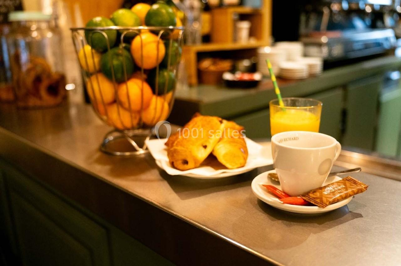 Petit-déjeuner sur un comptoir avec croissants, jus d'orange, café et corbeille de fruits en arrière-plan.