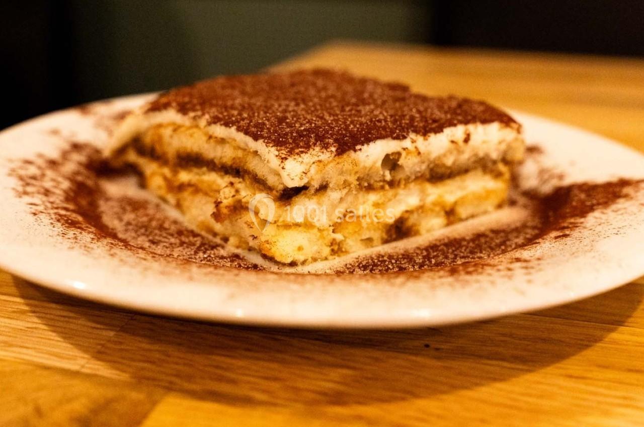 Part de tiramisu servie sur une assiette blanche, saupoudrée de cacao, posée sur une table en bois.
