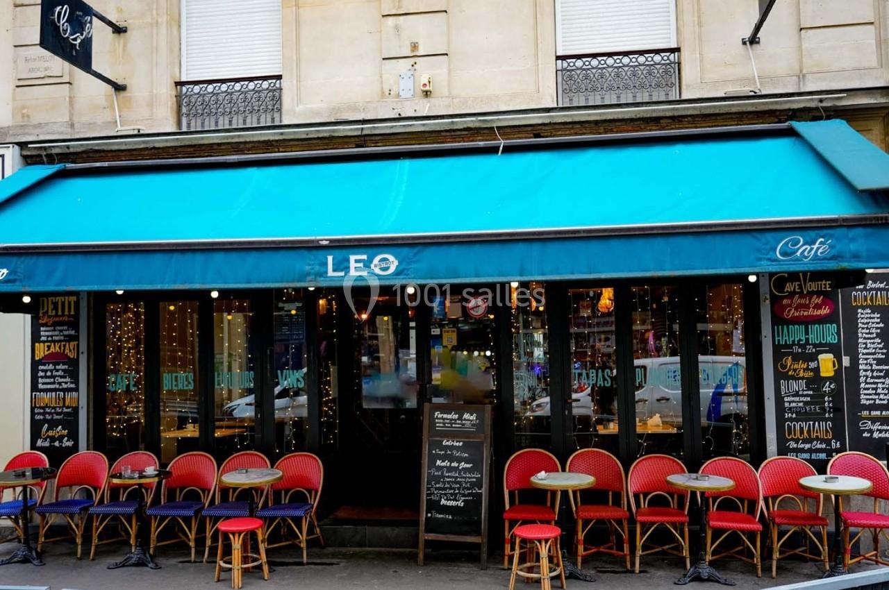Façade d'un café avec une terrasse composée de chaises rouges et de petites tables rondes sous un auvent bleu.