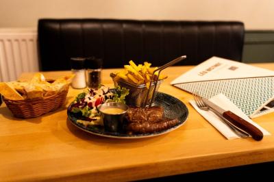 Façade d'un café-restaurant avec terrasse, chaises rouges et tables rondes, éclairage chaleureux à l'intérieur.