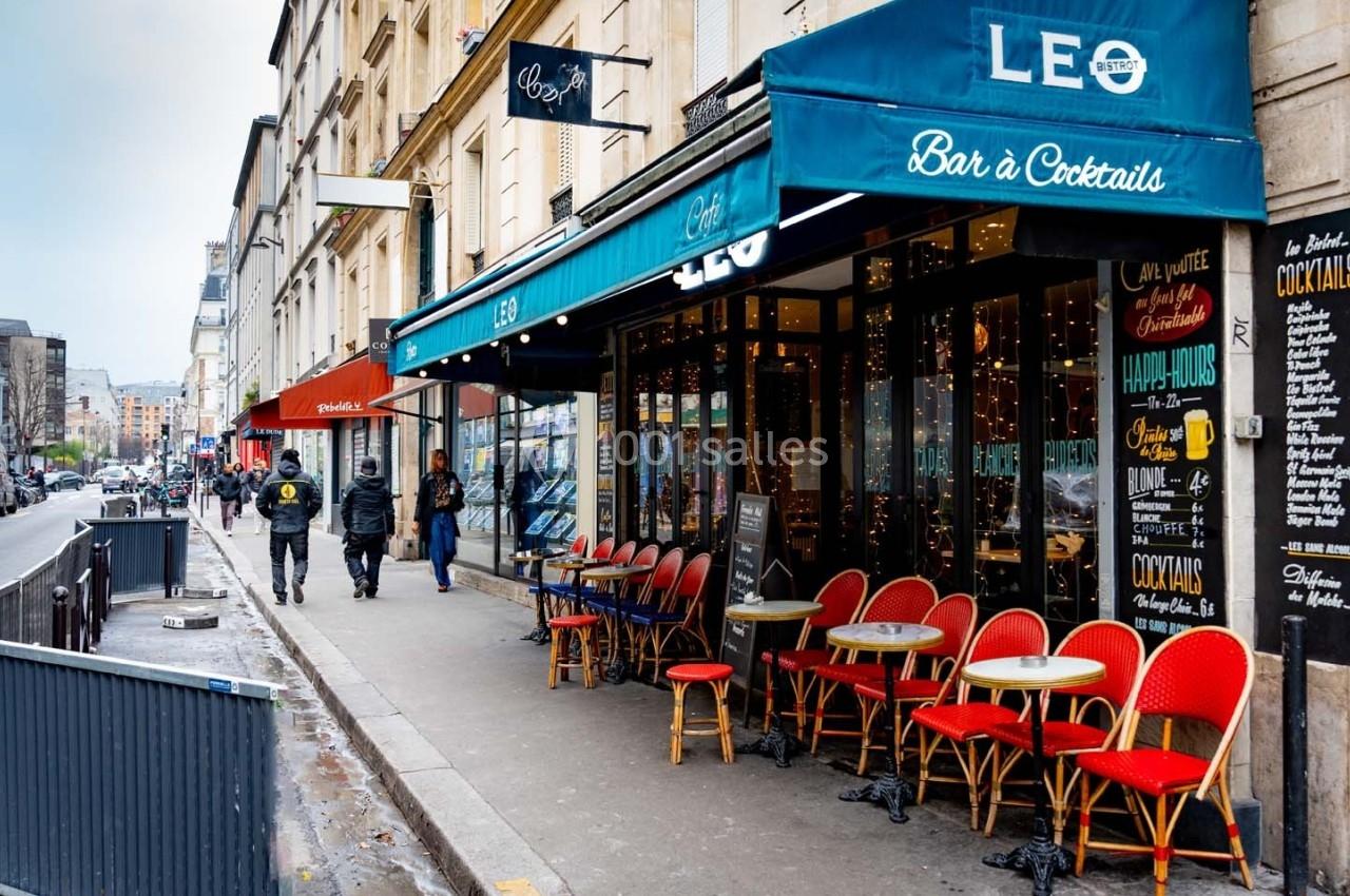 Façade d'un café avec terrasse et chaises rouges, situé dans une rue urbaine bordée de bâtiments.