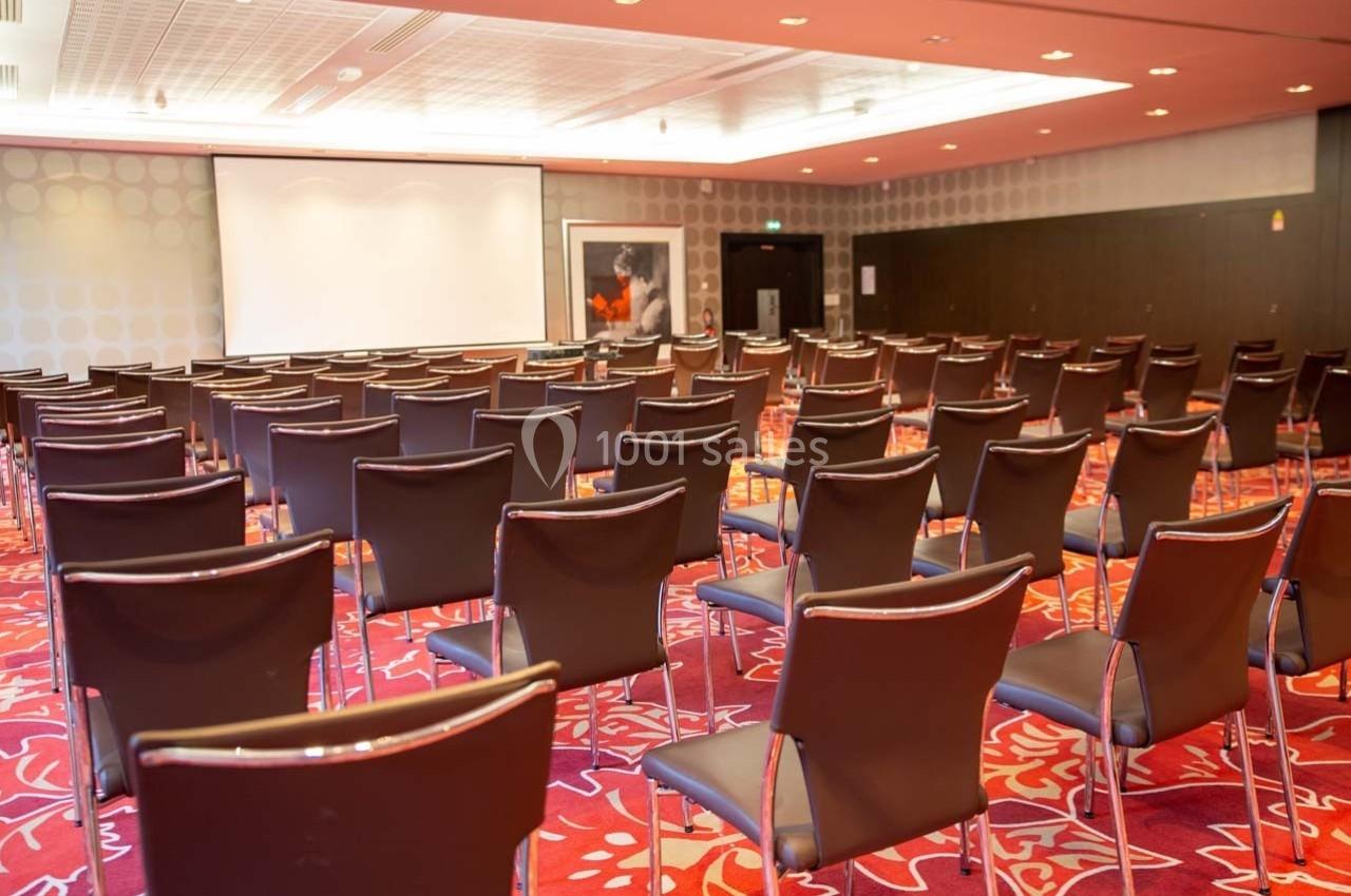 Salle de conférence vide avec rangées de chaises noires, écran de projection et moquette rouge à motifs.