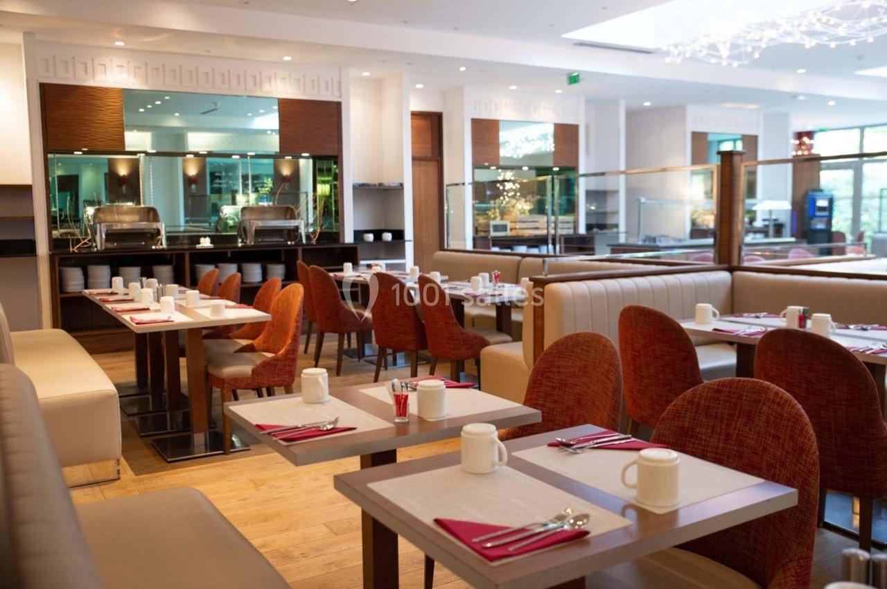 Salle de restaurant lumineuse avec tables dressées, chaises rembourrées et vue sur un buffet en arrière-plan.