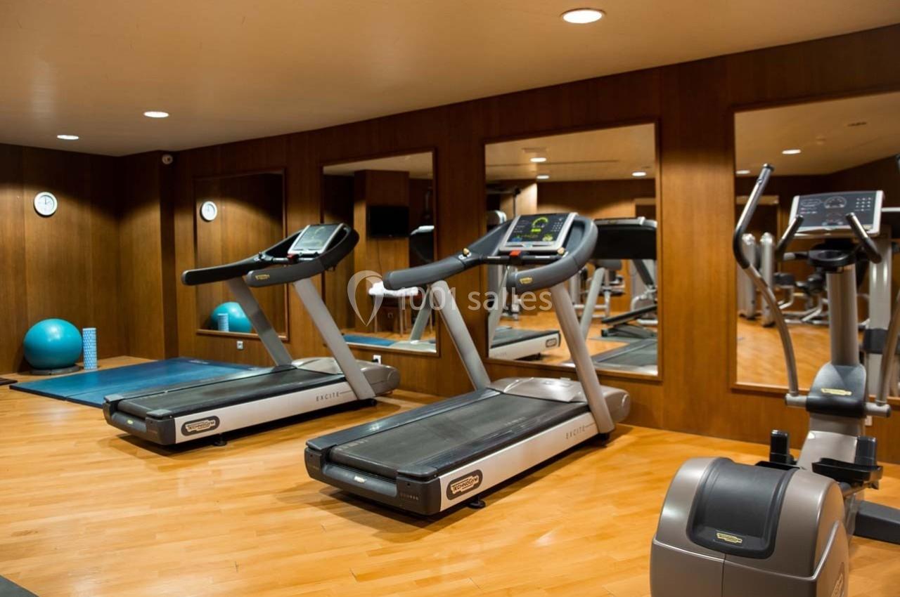 Salle de sport avec tapis de course, vélo d'exercice, miroir mural et équipements de fitness sur parquet clair.