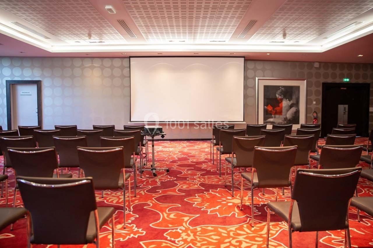 Salle de conférence vide avec des rangées de chaises, un écran de projection et un sol recouvert de moquette rouge.