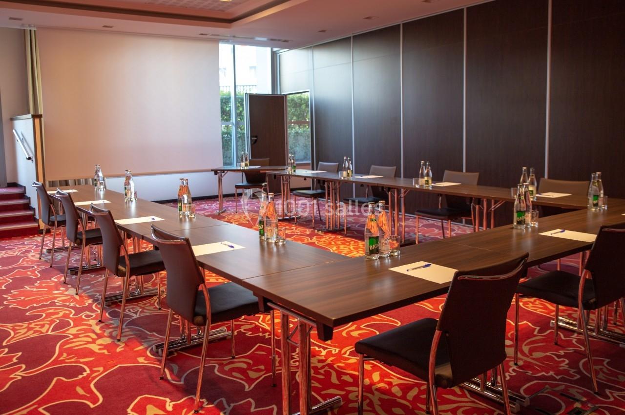 Salle de réunion avec tables disposées en U, chaises, bouteilles d'eau et blocs-notes, éclairée par une grande baie vitrée.