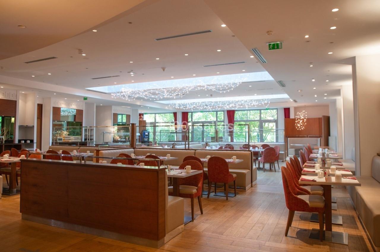 Salle de restaurant lumineuse avec tables dressées, chaises rouges et grande baie vitrée donnant sur un espace vert.