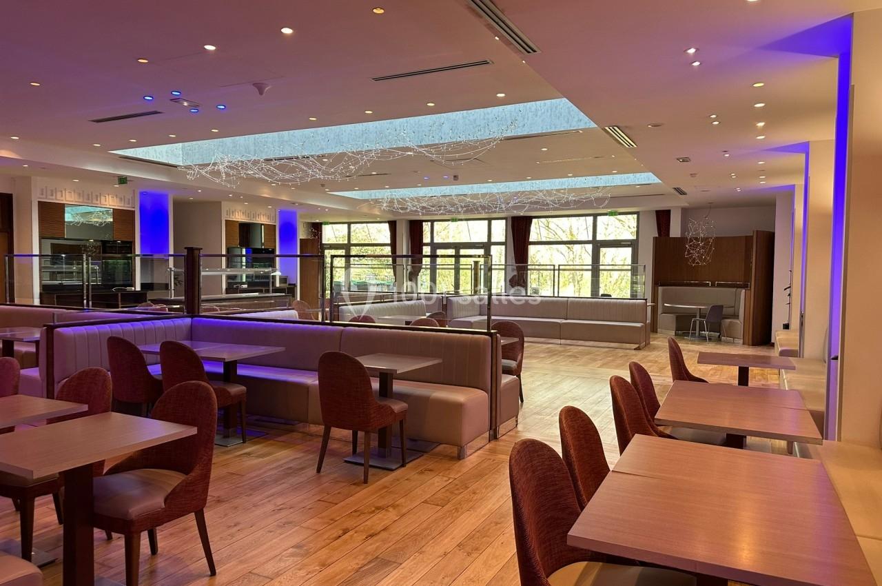 Salle de restaurant moderne avec tables en bois, chaises rembourrées, éclairage tamisé et grandes fenêtres donnant sur des…