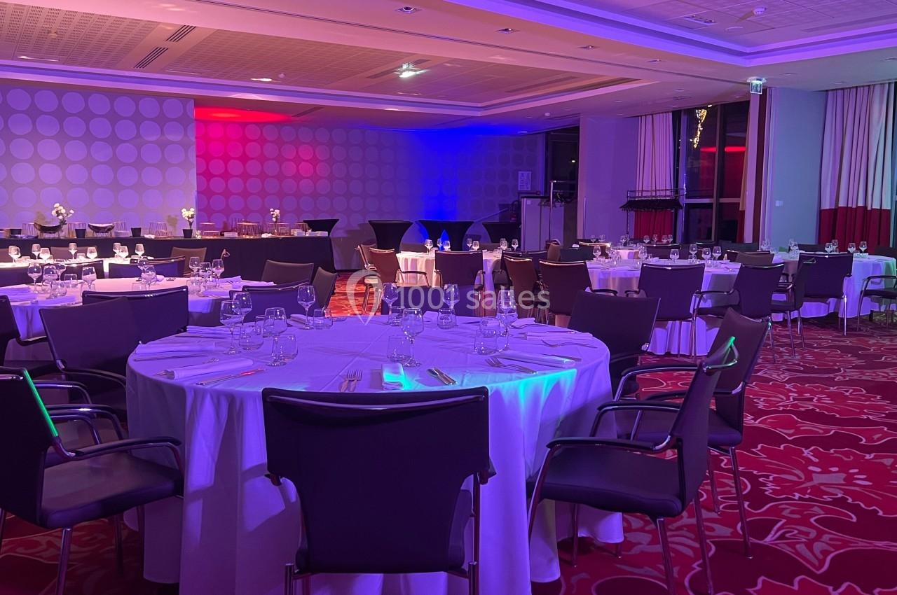 Salle de réception avec tables rondes dressées, nappes blanches, chaises noires et éclairage coloré.