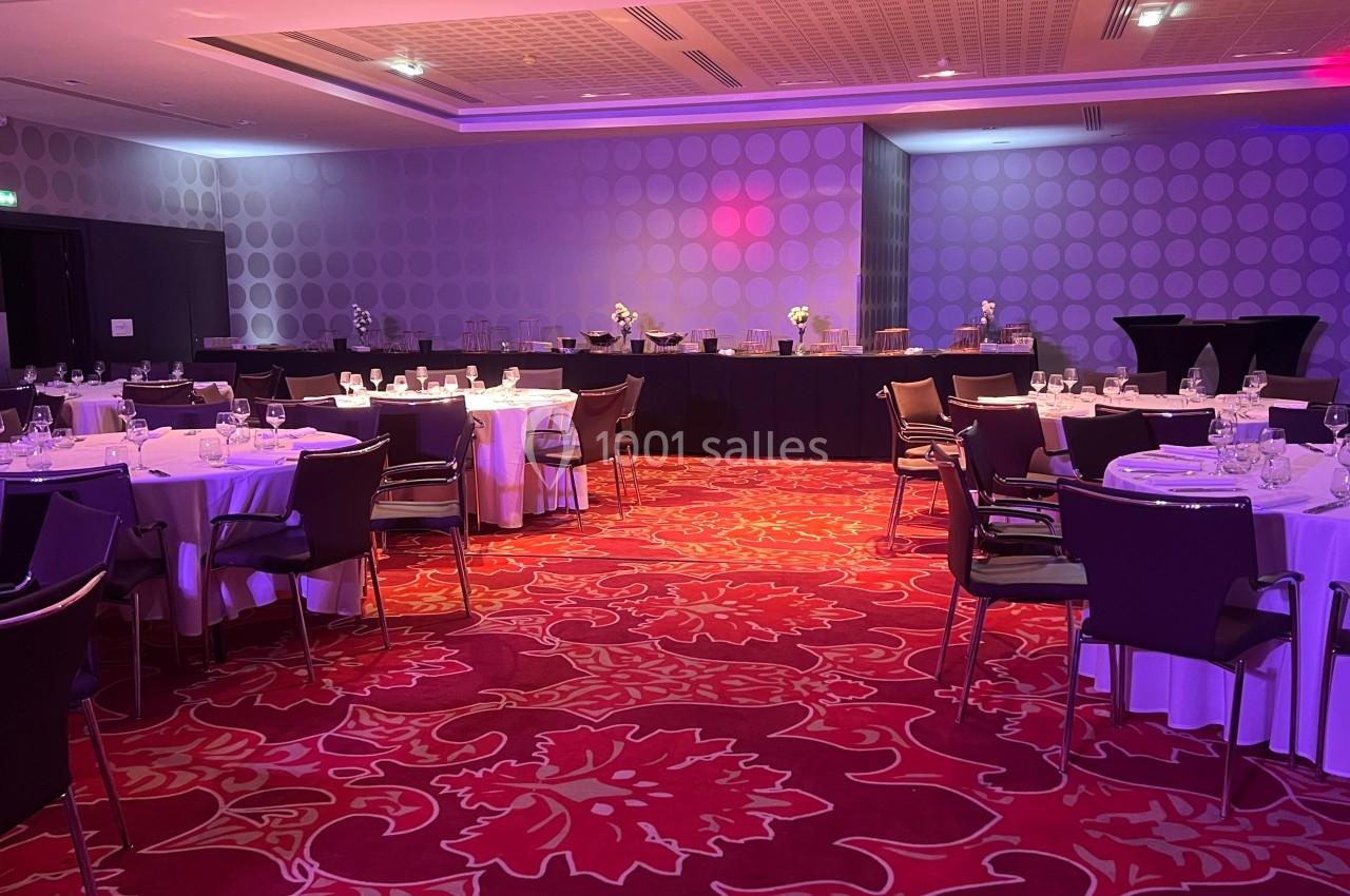 Salle de réception avec tables rondes dressées, nappes blanches, chaises modernes et éclairage tamisé.