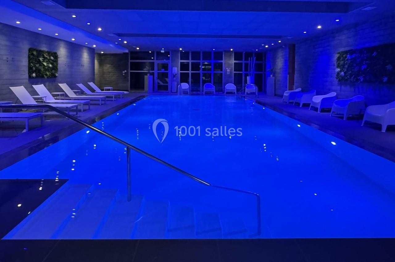 Piscine intérieure éclairée en bleu, entourée de chaises longues blanches et de murs décorés de plantes.