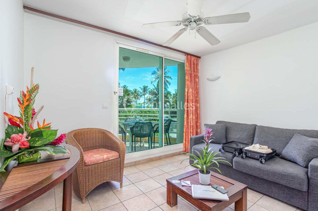 Salon lumineux avec canapé, fauteuil, table basse, bouquet de fleurs et vue sur un balcon donnant sur des palmiers.