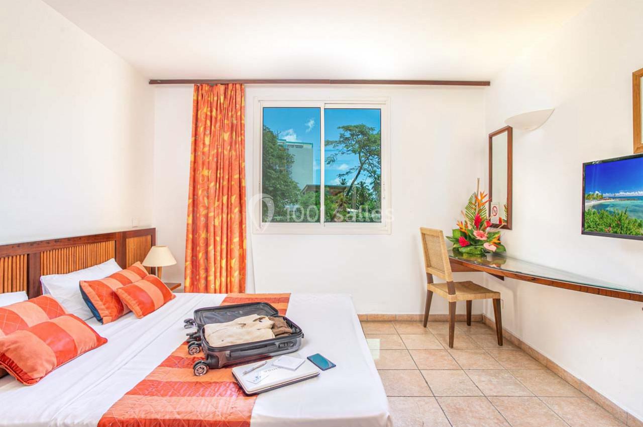 Chambre lumineuse avec lit double, valise ouverte, bureau, chaise, télévision, fenêtre avec vue sur la végétation.