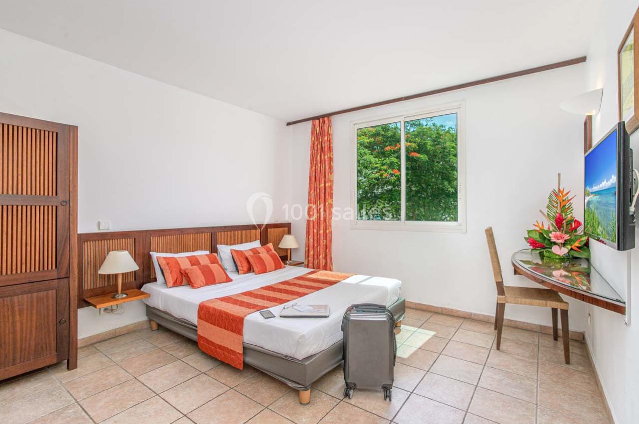 Chambre lumineuse avec lit double, valise au sol, bureau, armoire en bois et fenêtre donnant sur un jardin verdoyant.