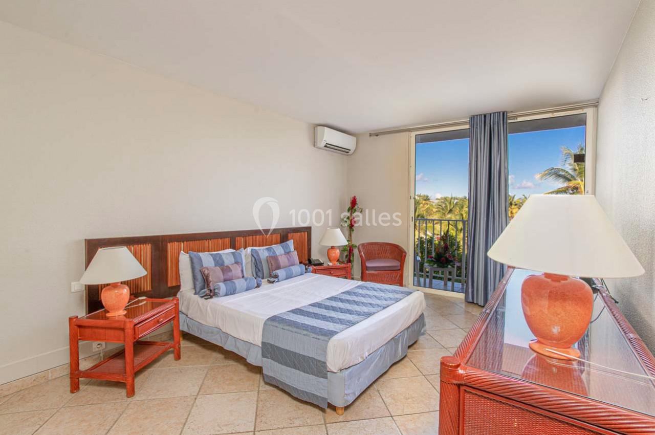 Chambre lumineuse avec lit double, mobilier en bois, lampes, balcon donnant sur un paysage tropical et ciel bleu.