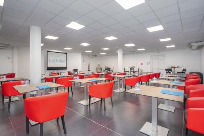 Salle de réunion avec tables disposées en U, chaises bleues et rouges, éclairage au plafond et sol carrelé beige.