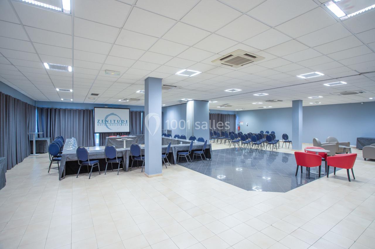 Salle spacieuse avec tables et chaises disposées pour une réunion ou un événement, éclairée par des lumières au plafond.