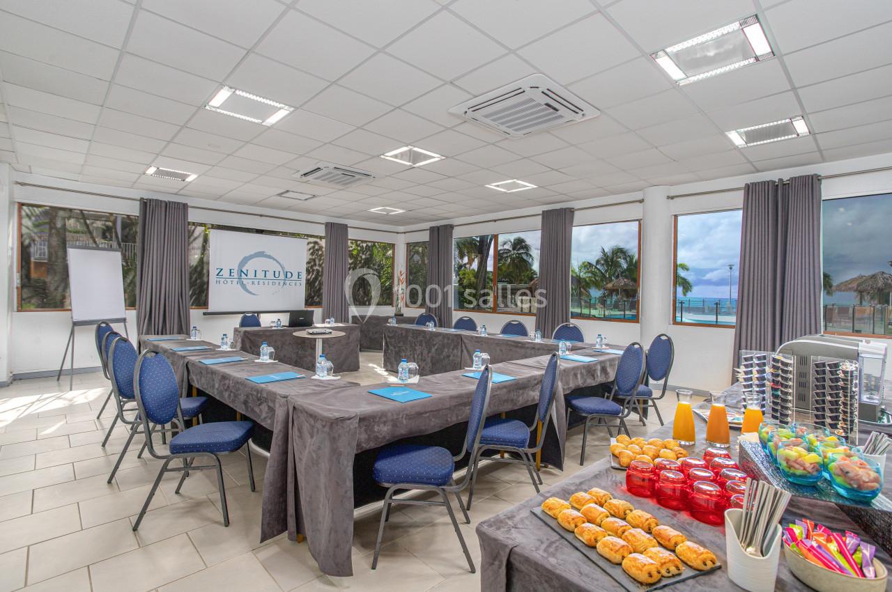 Salle de réunion lumineuse avec tables disposées en U, boissons, viennoiseries et vue sur la mer à travers les fenêtres.