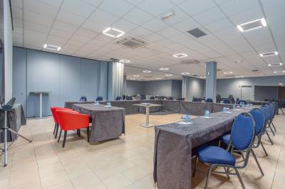 Salle de réunion avec tables disposées en U, chaises bleues et rouges, éclairage au plafond et sol carrelé beige.