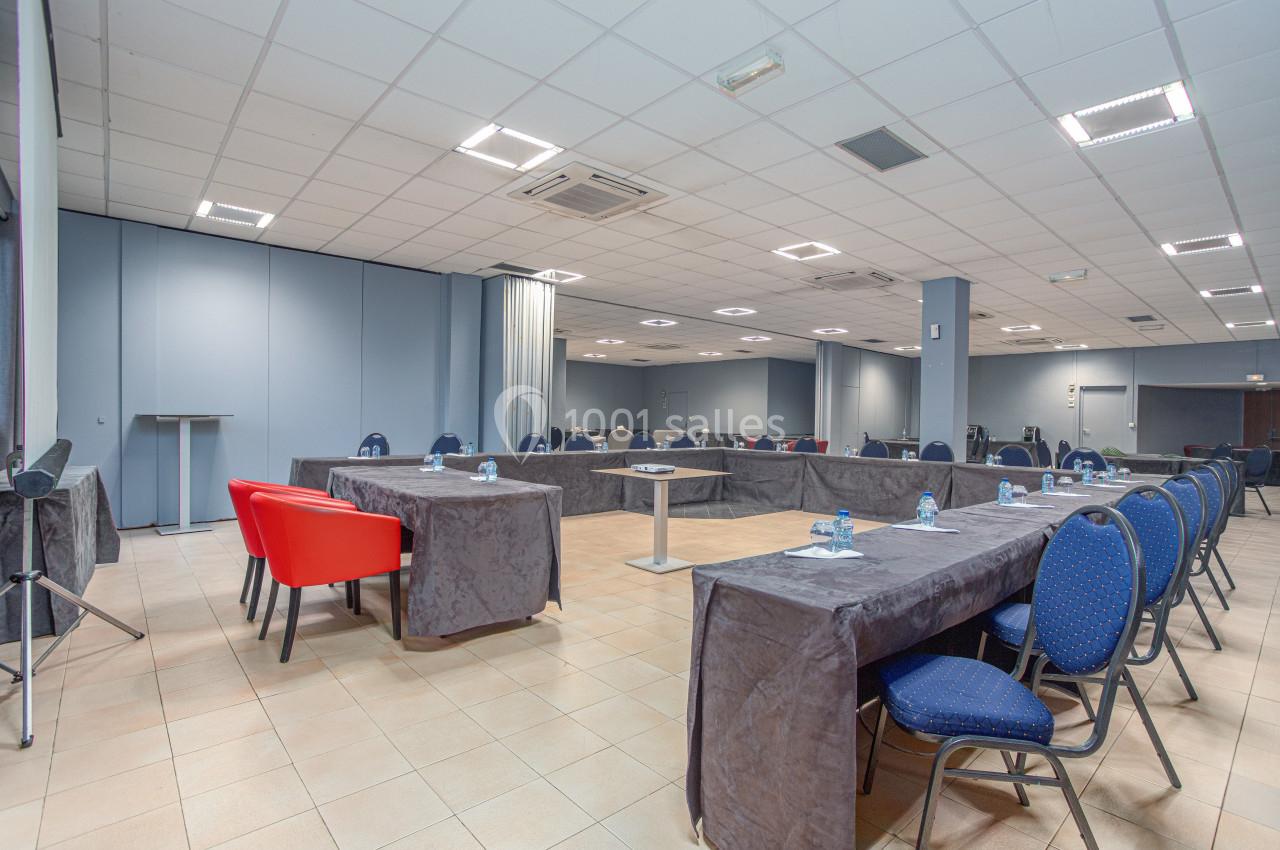 Salle de réunion avec tables disposées en U, chaises bleues et rouges, éclairage au plafond et sol carrelé beige.