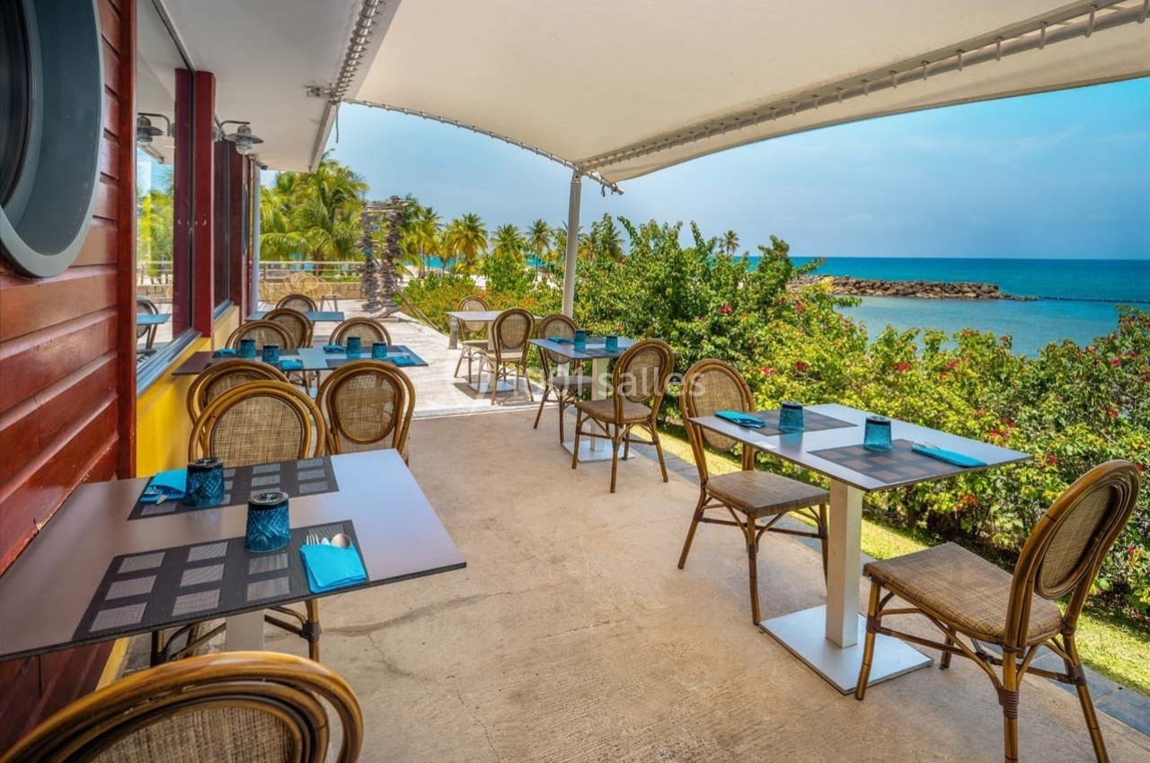 Terrasse d'un restaurant avec tables et chaises en osier, vue sur la mer, entourée de végétation tropicale.