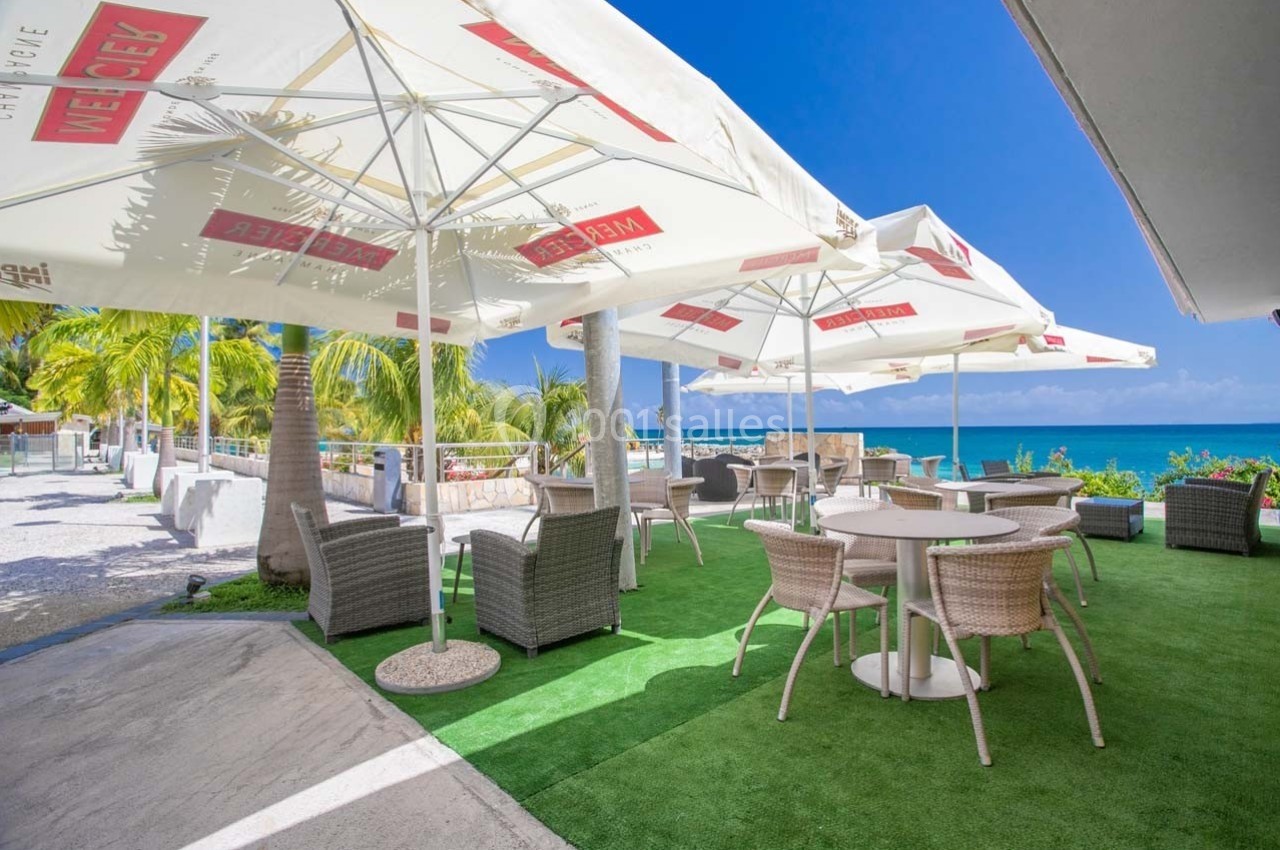 Terrasse avec tables et chaises en rotin sous des parasols blancs, vue sur la mer et palmiers en arrière-plan.