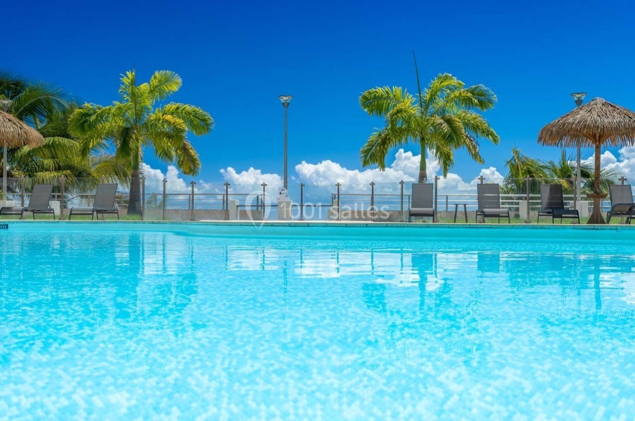 Piscine extérieure avec eau turquoise, entourée de palmiers, chaises longues et ciel bleu en arrière-plan.