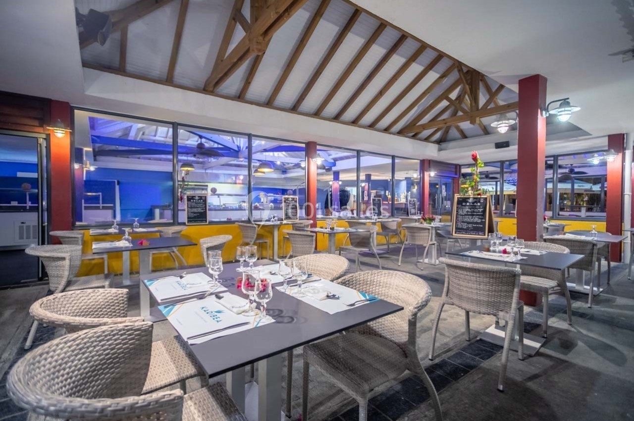 Salle de restaurant avec tables dressées, chaises en rotin, toit en bois apparent et vue sur un espace cuisine vitré.