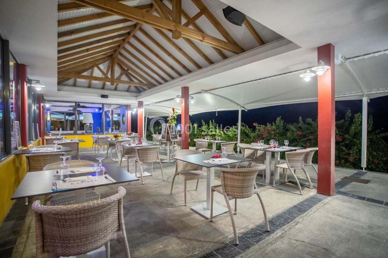 Terrasse couverte d'un restaurant avec tables dressées, chaises en rotin et vue sur un jardin éclairé en soirée.