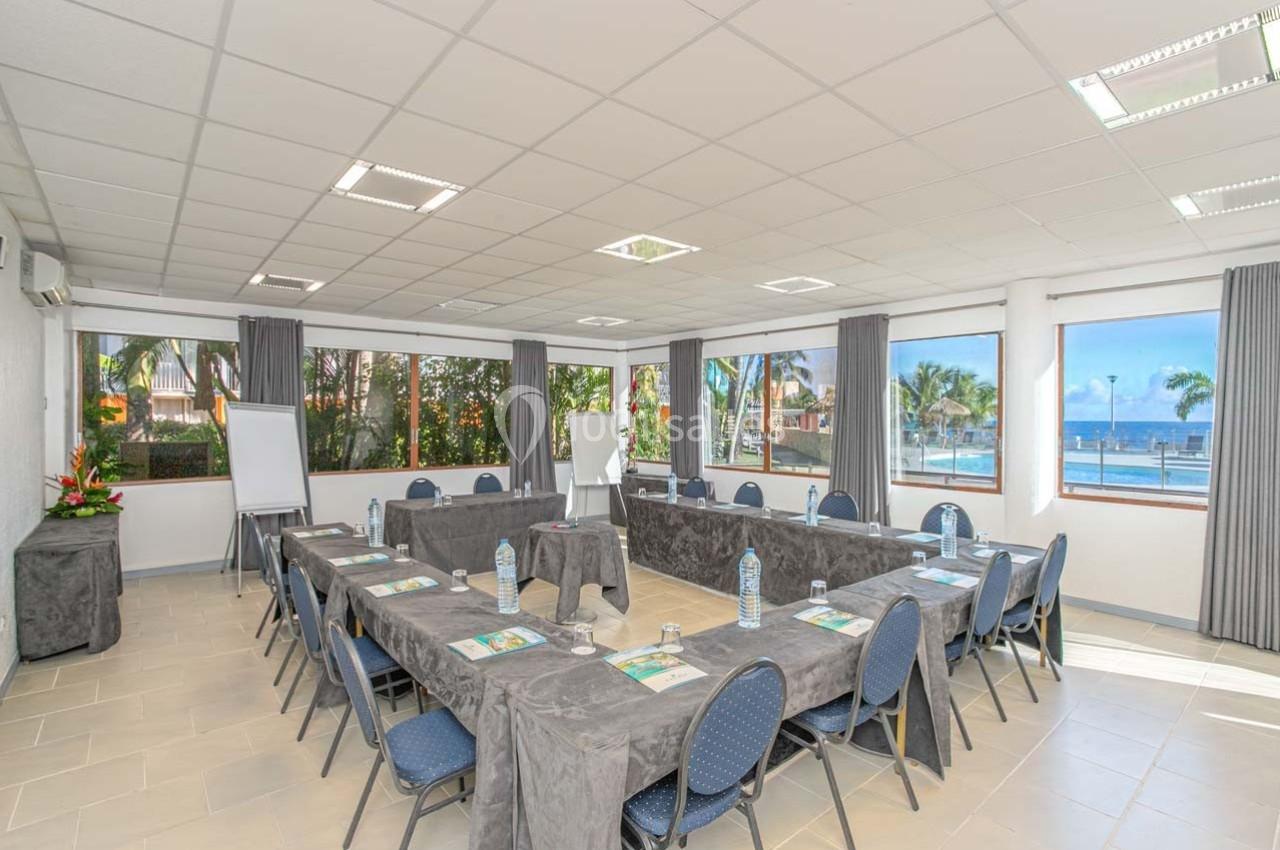Salle de réunion lumineuse avec tables en U, chaises, paperboard et vue sur un paysage tropical avec la mer.