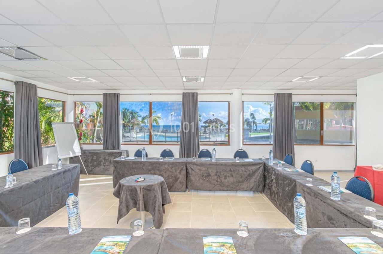 Salle de réunion lumineuse avec vue sur la mer, tables disposées en U, bouteilles d'eau et documents présents.