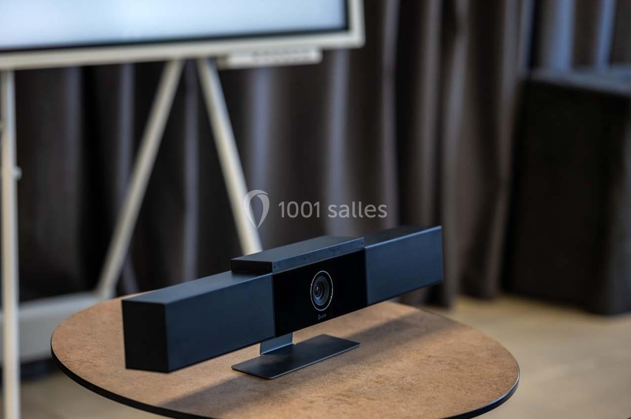 Caméra de visioconférence noire posée sur une table ronde devant un écran et des rideaux sombres.