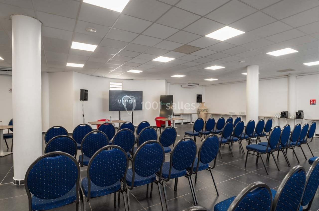 Salle de conférence avec des chaises bleues disposées face à un écran et un tableau dans un espace lumineux.