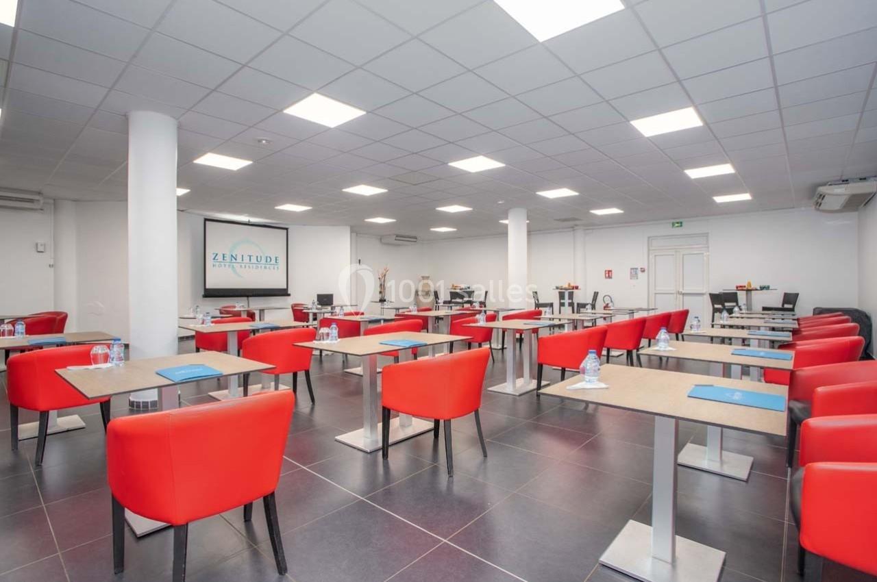 Salle de réunion lumineuse avec des tables individuelles, chaises rouges et matériel de conférence disposé.