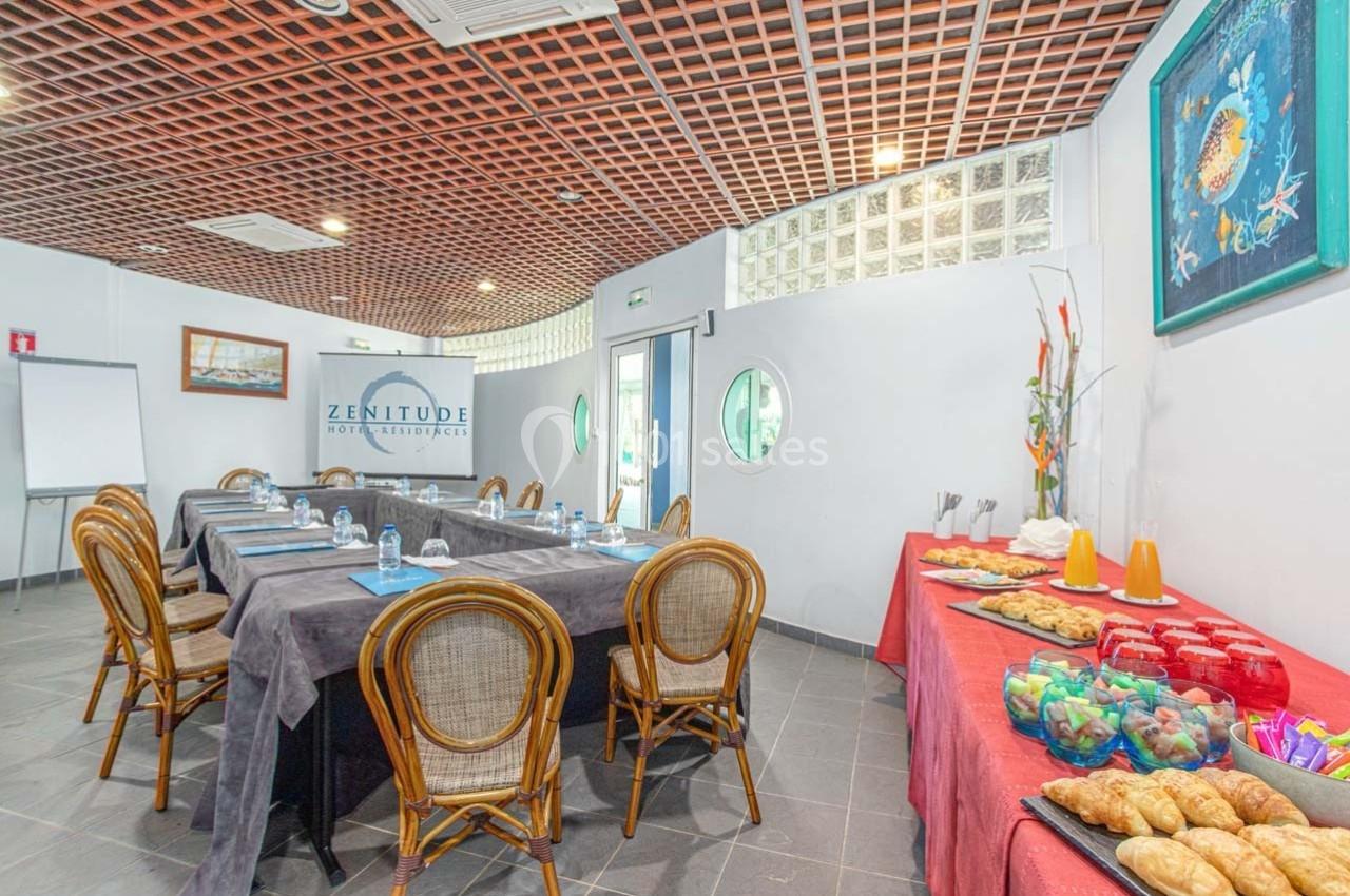 Salle de réunion avec tables dressées, chaises en rotin, et buffet comprenant viennoiseries, boissons et fruits.