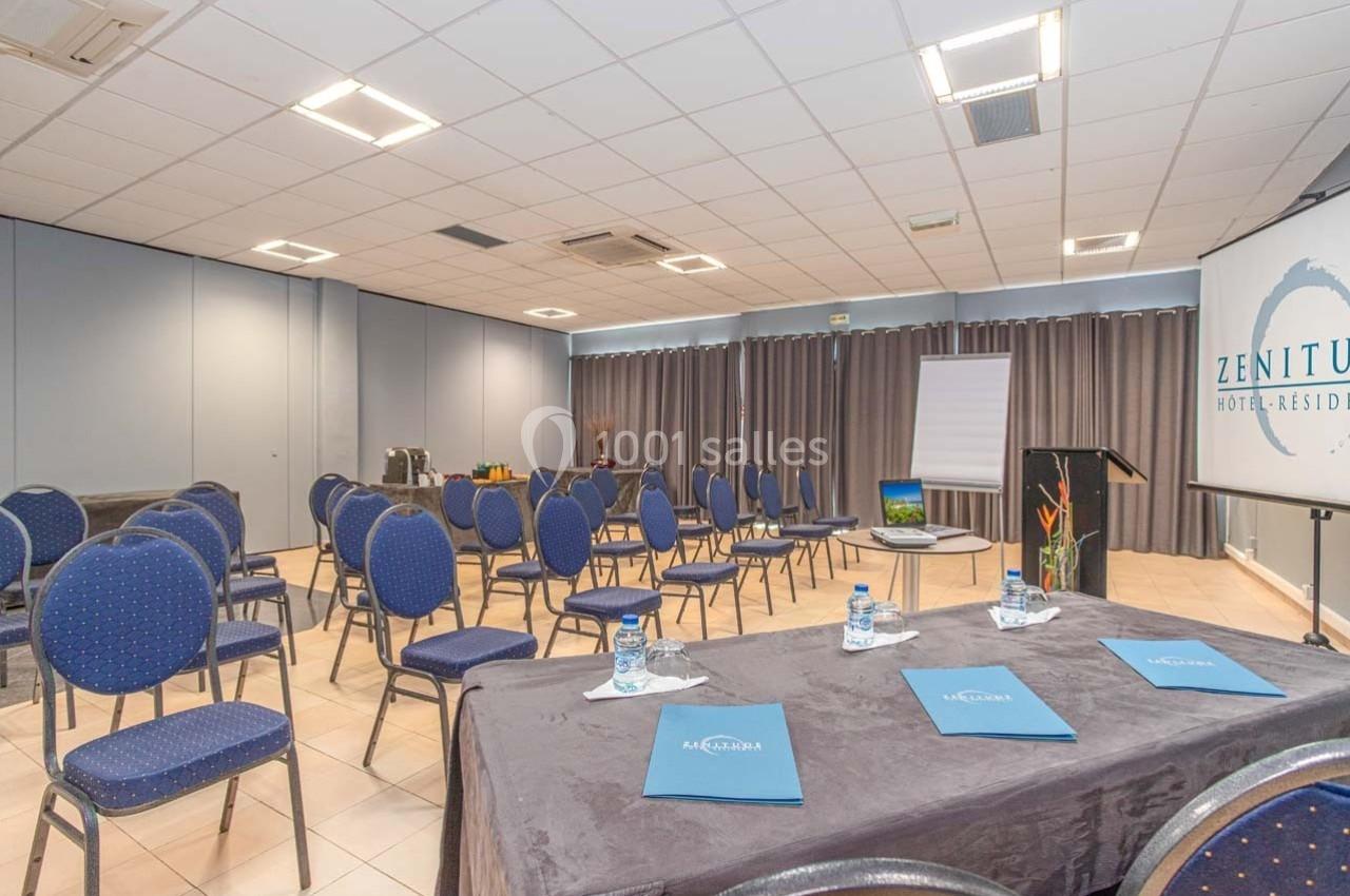 Salle de conférence lumineuse avec chaises disposées en rangées, table équipée et écran de projection.