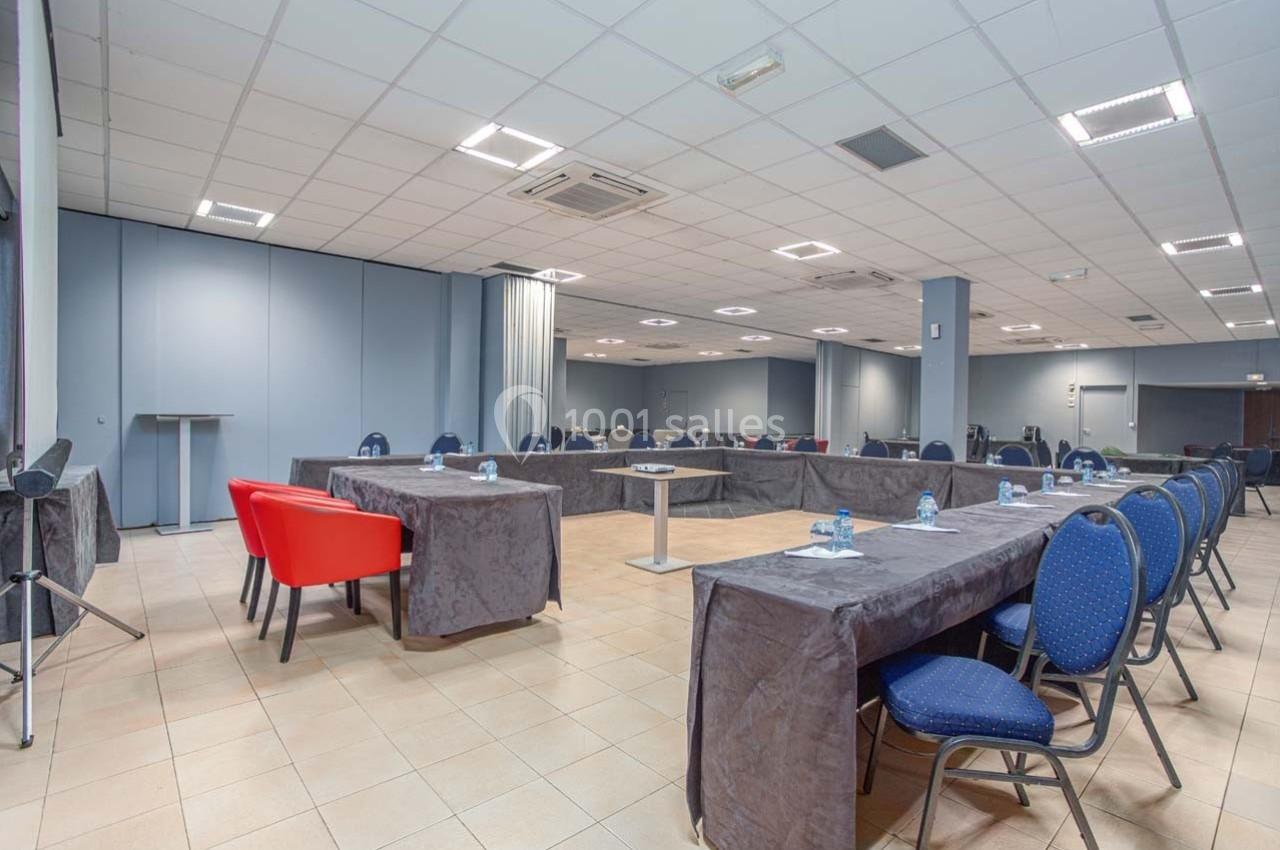 Salle de réunion lumineuse avec tables en U couvertes de nappes grises, chaises bleues et rouges, et bouteilles d'eau.