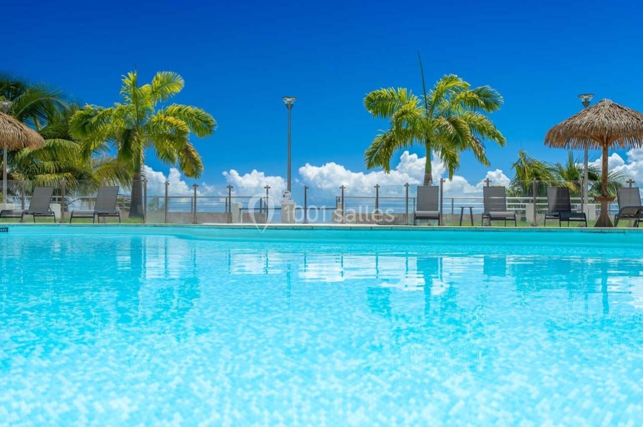 Piscine extérieure avec eau turquoise, entourée de palmiers et de chaises longues sous un ciel bleu dégagé.