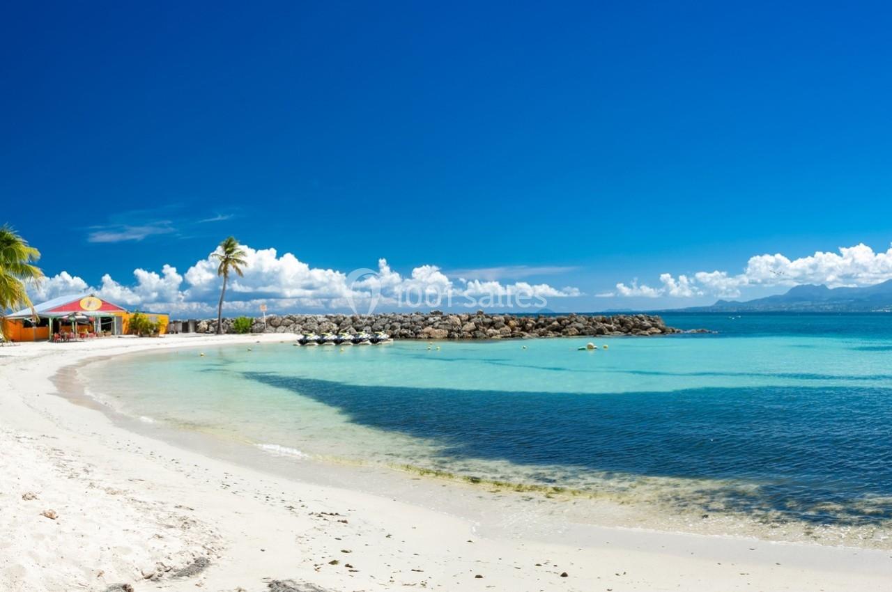 Plage de sable blanc bordée de palmiers, mer turquoise calme et ciel bleu dégagé avec quelques nuages.