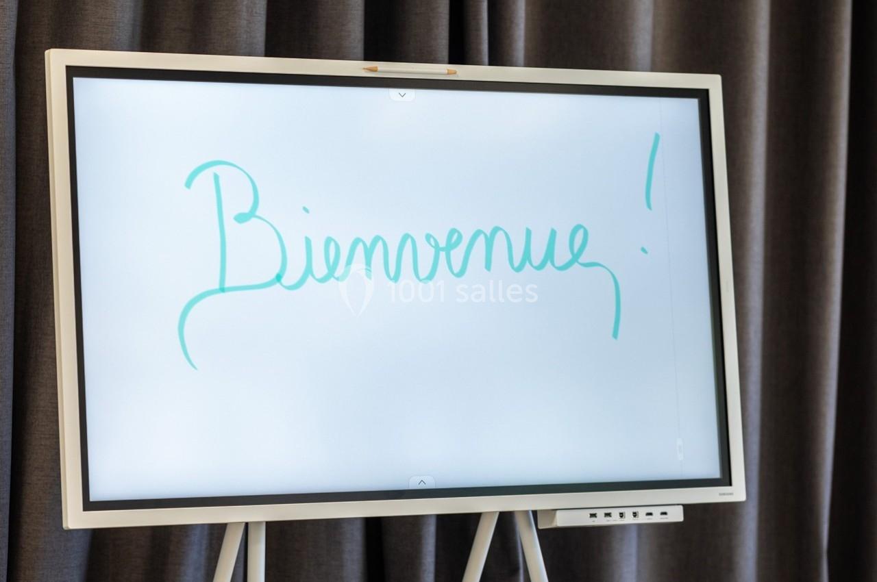 Écran blanc interactif affichant le mot ’Bienvenue !’ écrit en lettres vertes, devant un rideau sombre.